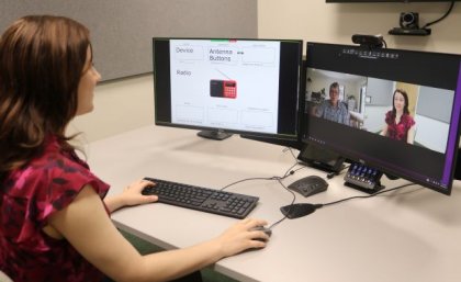 A woman sits at a desk looking at two monitors while she conducts a Zoom call with her face and a man's shown on one of the screens.